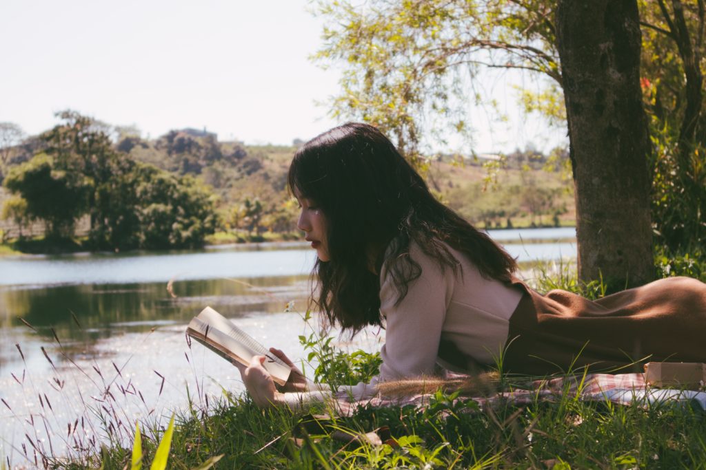 Beautiful Asian woman reads a book on a grassy riverbank.